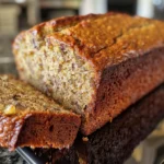 A loaf of 3-ingredient banana bread is sliced on a wooden cutting board, with two ripe bananas and eggshells in the background.