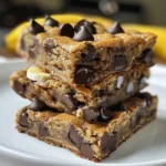 Four chocolate chip banana bread bars arranged on a wooden cutting board, next to a bowl of chocolate chips and a banana.